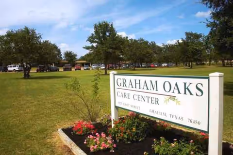 Outdoor scene with Graham Oaks Care Center sign, grassy lawn, trees, and parked cars in background.
