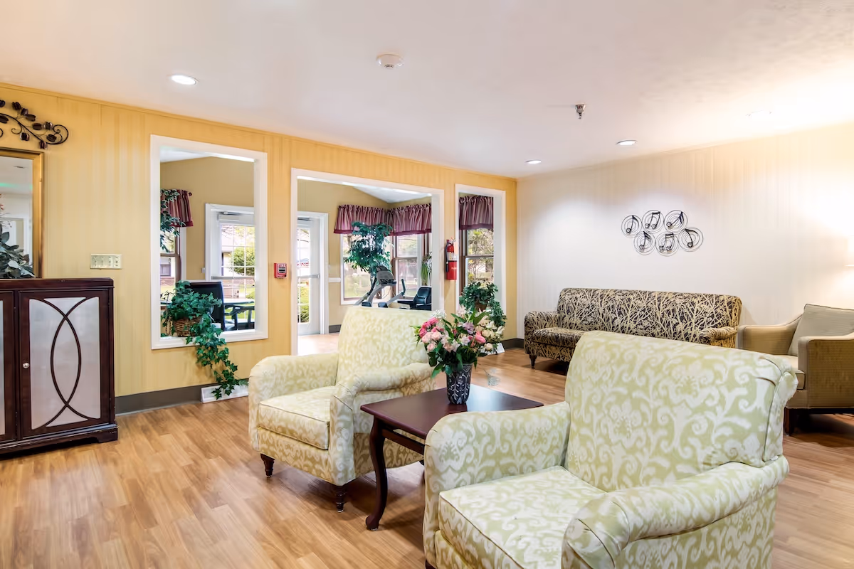 A cozy living room area with two patterned armchairs and a small wooden coffee table with a vase of flowers. In the background, there is a patterned sofa, an armchair, and a wall decoration. The room has wooden flooring, yellow and white walls, and windows with purple valances letting in natural light.