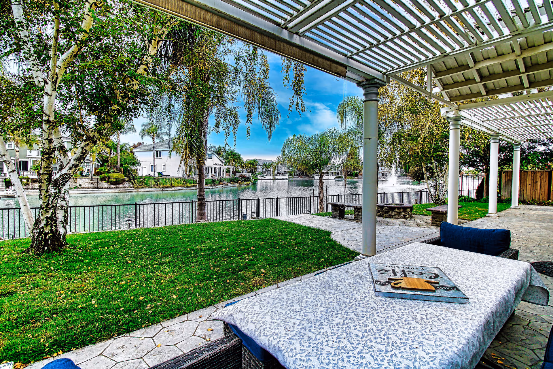 A covered outdoor patio area with a table covered by a patterned tablecloth and a book on top. The patio overlooks a green lawn, a black metal fence, and a serene lake with palm trees and houses in the background under a partly cloudy blue sky.