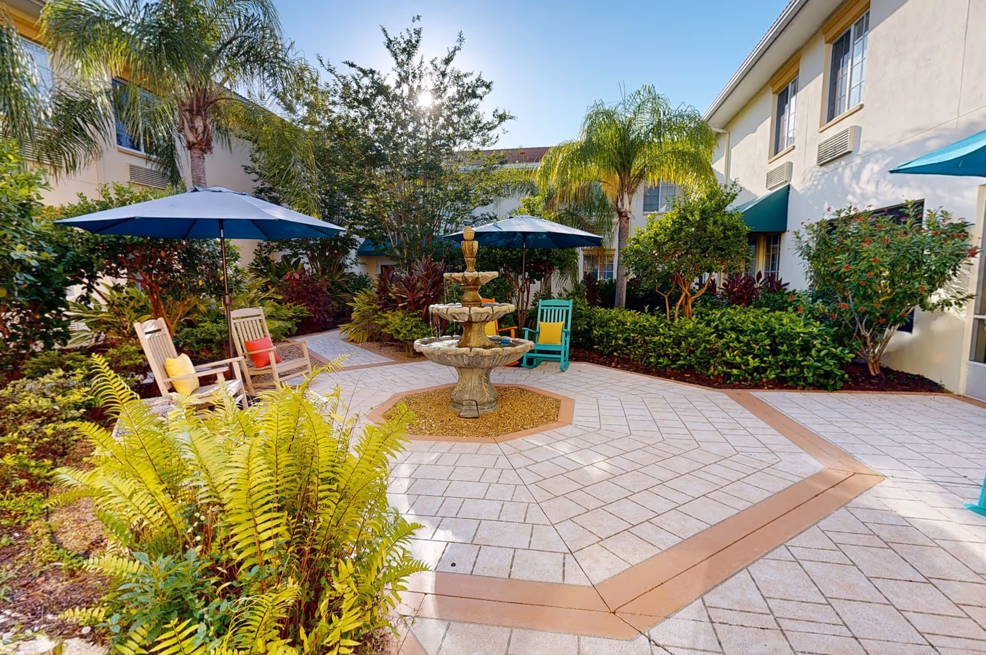 Outdoor courtyard area with a central tiered stone fountain surrounded by tiled flooring. There are several wooden chairs with colorful cushions and blue umbrellas providing shade. The courtyard is bordered by lush green plants, palm trees, and shrubs, with a two-story building in the background under a clear blue sky.