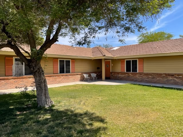 Single-story building front with a grassy courtyard, a tree, and a small patio area with two chairs in front of beige siding and brick facade.