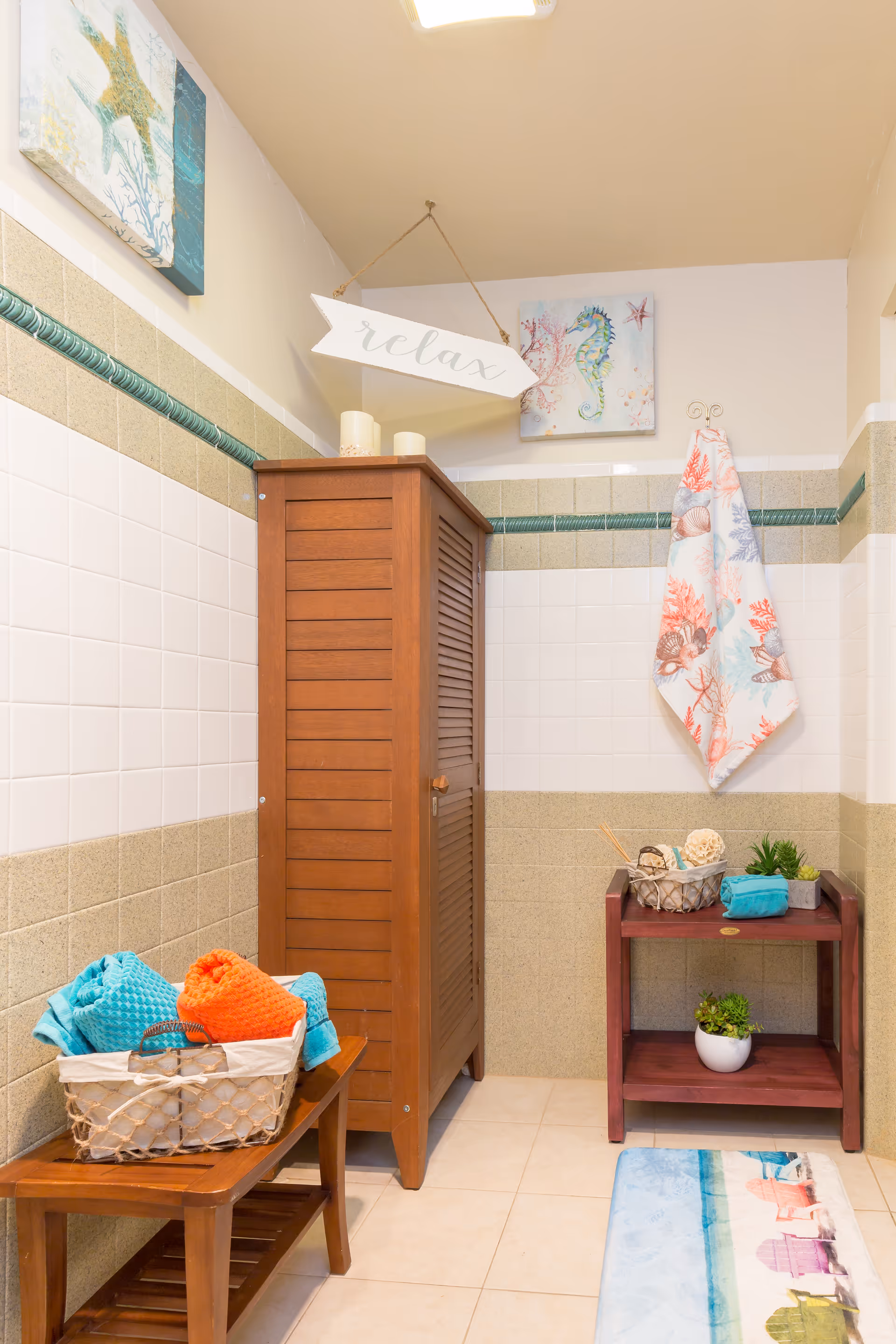 Tiled bathroom interior featuring a wooden cabinet, a bench with colorful towels, a small side table with decor, and a hanging "relax" sign.