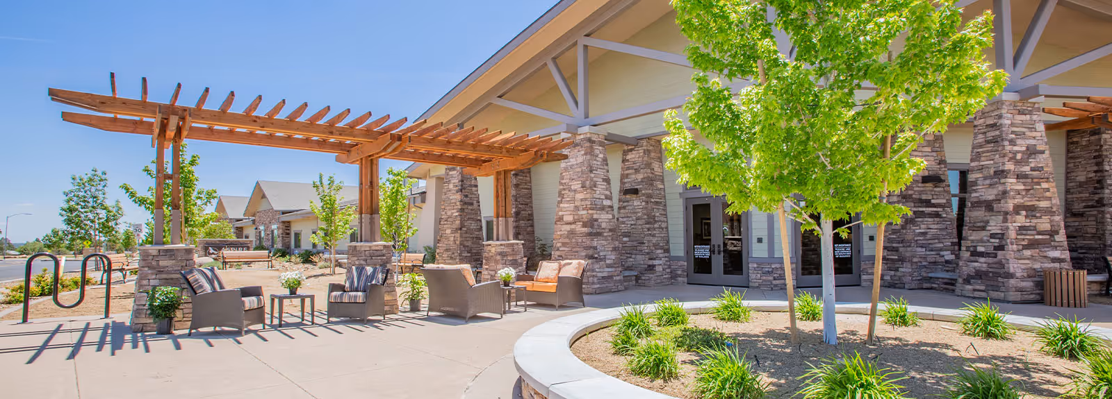 Outdoor seating area at Aspire Transitional Care featuring wicker chairs and small tables under a wooden pergola, with stone pillars and a building entrance in the background. There are green trees and plants around the paved area under a clear blue sky.