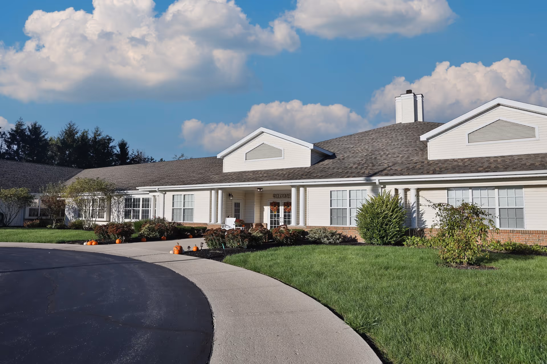 Exterior view of a single-story senior living facility building with white siding and a gray shingled roof under a partly cloudy blue sky. The entrance is centered with white columns and a small porch area, surrounded by green grass, shrubs, and small pumpkins placed along the walkway.