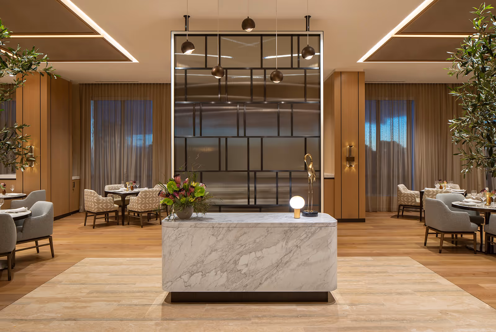 A modern and elegant dining area in Ellore Senior Living featuring a central marble reception or display table with a flower arrangement and a decorative lamp. The room has wooden floors, beige walls with vertical paneling, large windows with sheer curtains, and several dining tables with upholstered chairs arranged around the space. A large decorative metal and glass partition with hanging pendant lights is positioned behind the marble table.