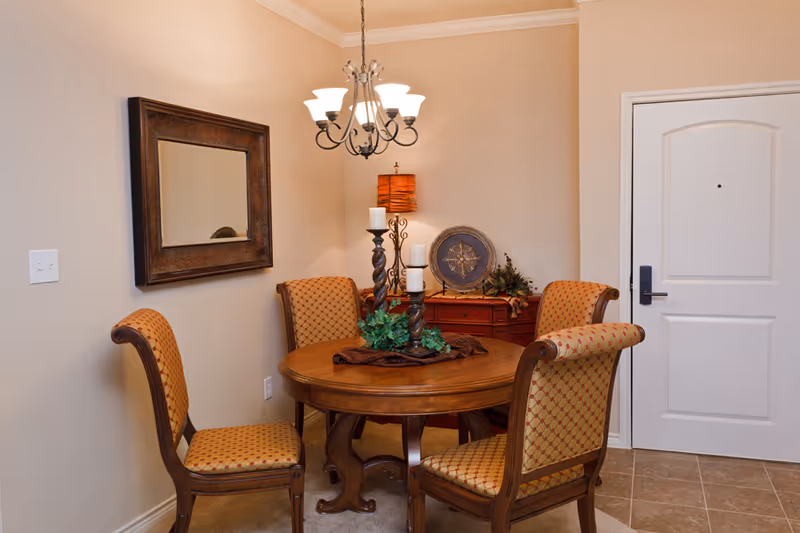 A cozy dining area with a round wooden table surrounded by four upholstered chairs with patterned fabric. On the table, there is a decorative centerpiece with candles and greenery. A chandelier with multiple lights hangs above the table. A large framed mirror is mounted on the wall to the left, and a white door with a black handle is visible on the right. In the corner, there is a wooden sideboard with decorative items including a lamp, a plate, and some greenery.