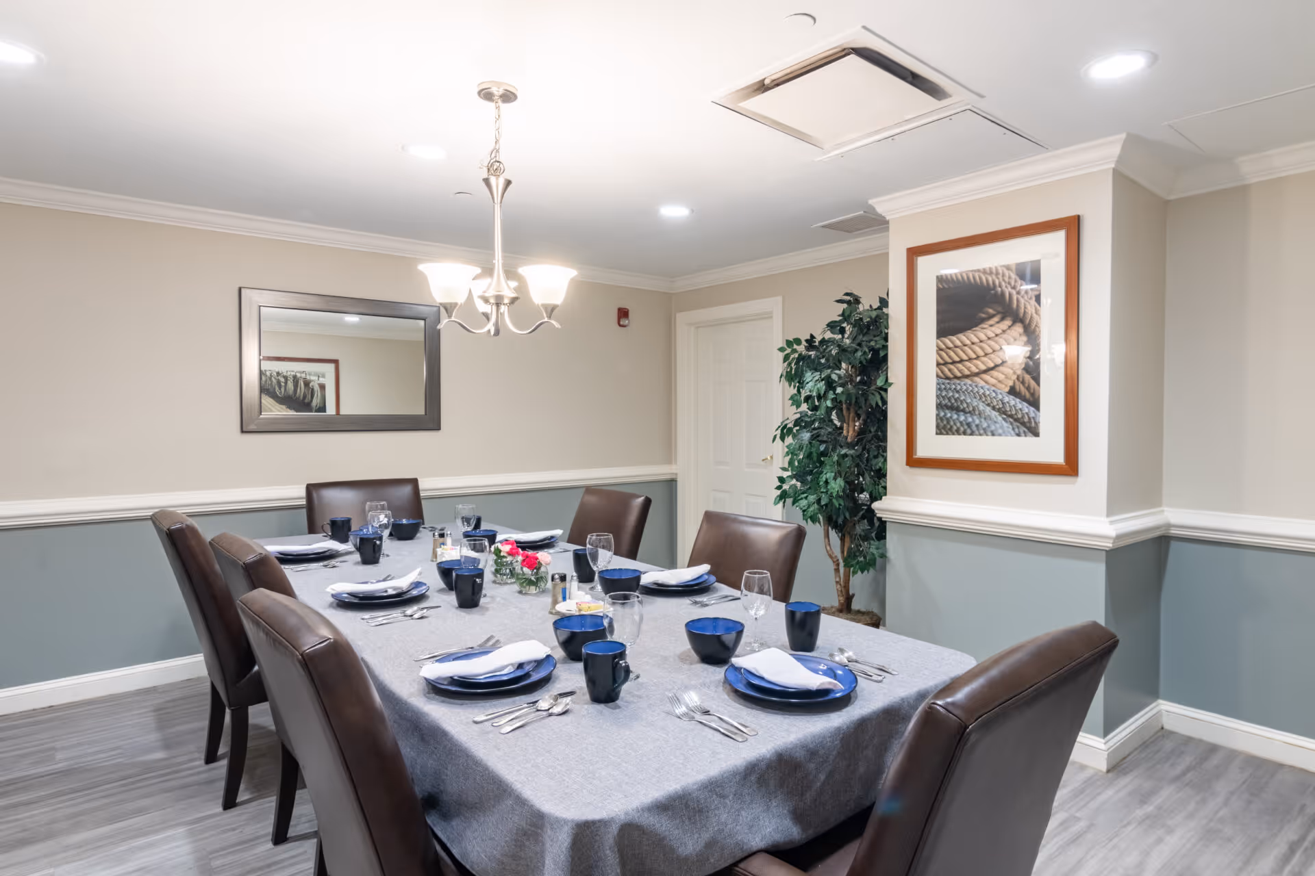 A dining room with a rectangular table covered with a gray tablecloth set for eight people. The table settings include blue plates, bowls, mugs, silverware, and napkins. The room has light-colored walls with a chair rail and a framed picture of coiled ropes on one wall. There is a large mirror on another wall and a potted plant in the corner. A chandelier with five lights hangs above the table.
