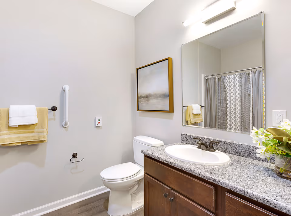 A clean and modern bathroom featuring a white toilet, a granite countertop with a built-in sink, a large mirror above the sink, a towel rack with yellow and white towels, a grab bar on the wall, a piece of framed artwork, and a shower curtain with a geometric pattern.