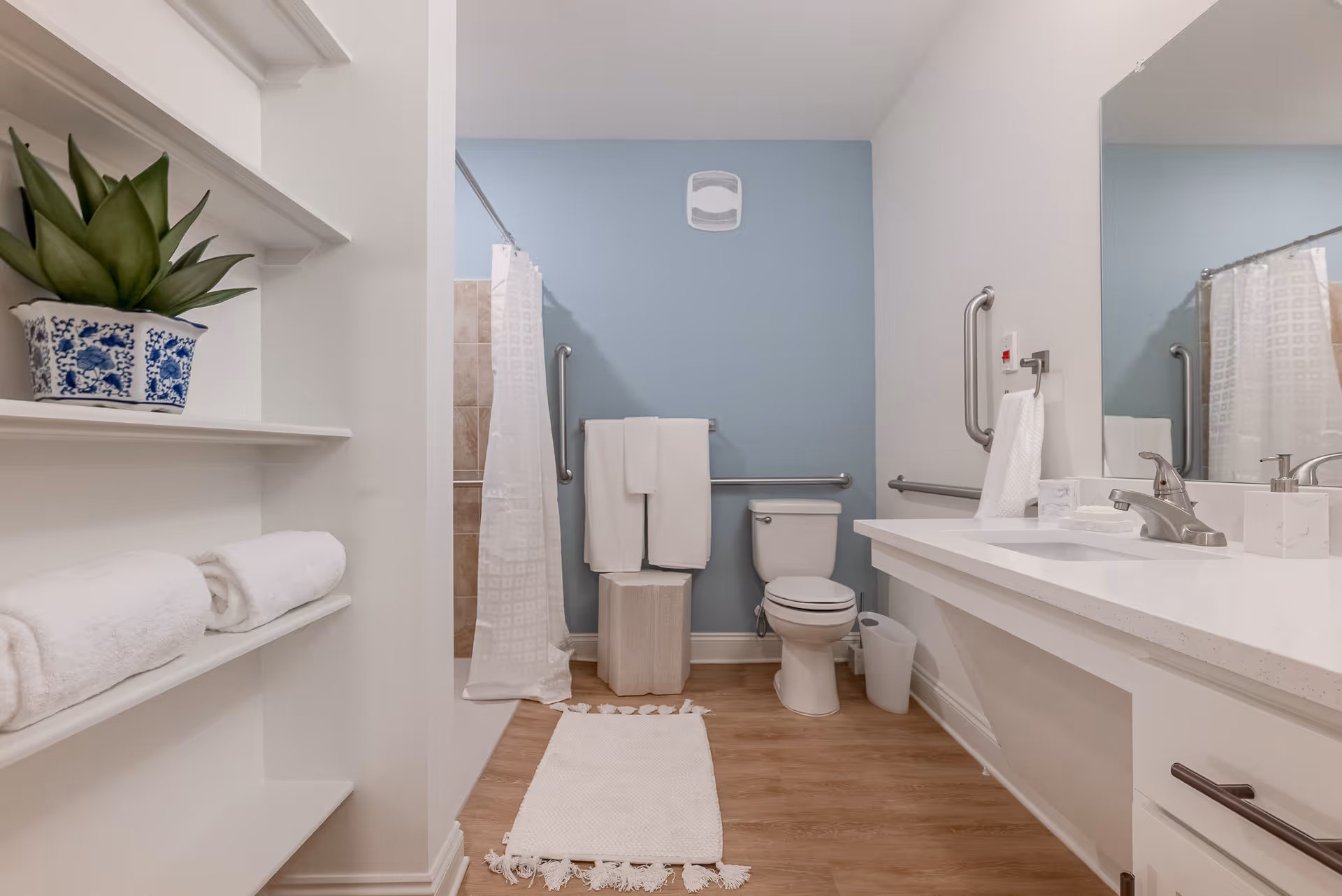 A clean and modern bathroom with a white countertop and sink on the right, a toilet in the center against a light blue wall, and a shower with a white curtain on the left. There are grab bars near the toilet and shower for accessibility. White towels are neatly folded on shelves and hanging on a towel rack. A small plant in a blue and white pot is placed on the shelves. The floor is wood-style, and a white rug is placed in front of the shower and toilet.