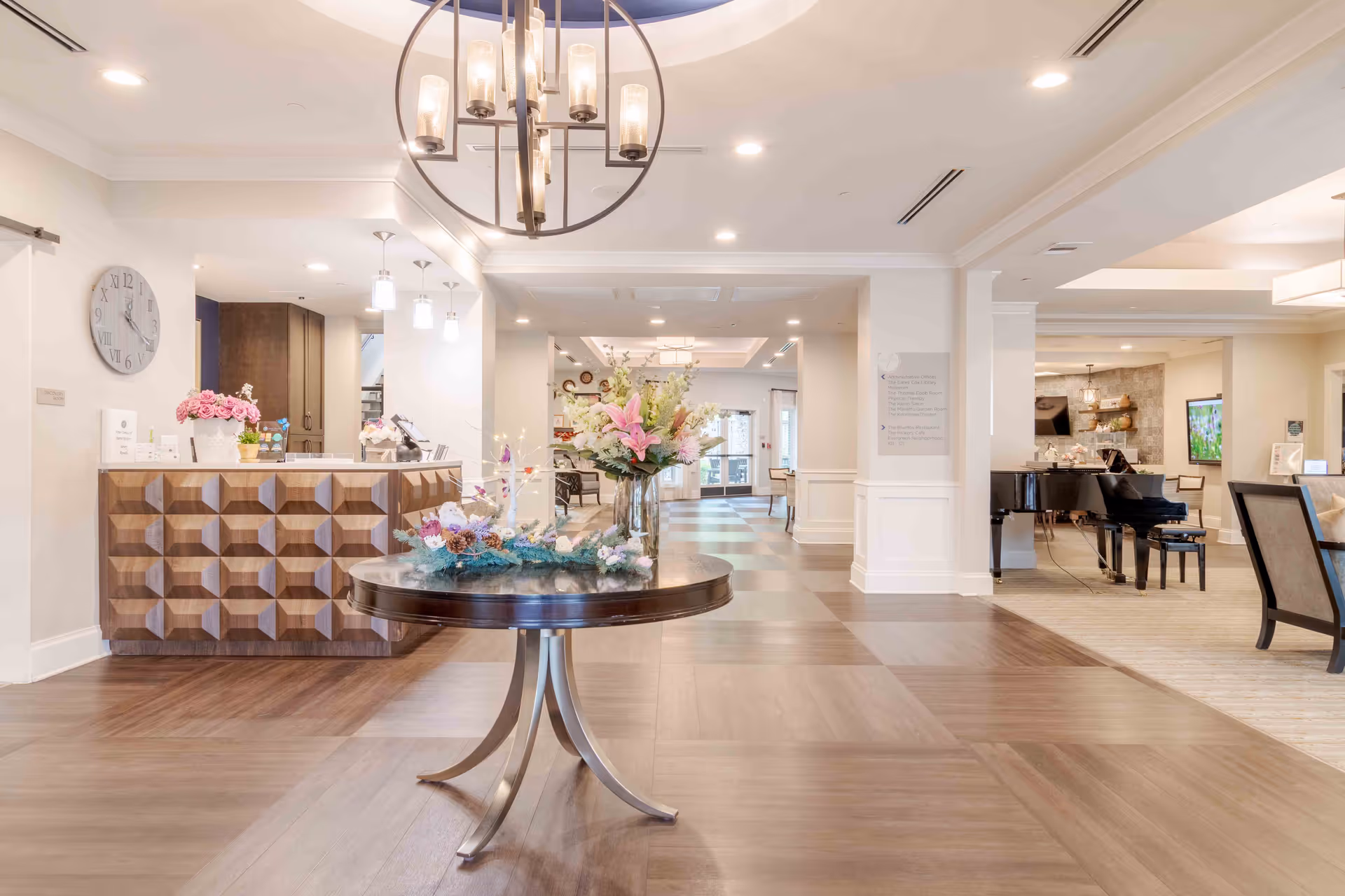 Bright senior living lobby with a round table floral centerpiece, reception desk on the left and a grand piano seating area on the right.