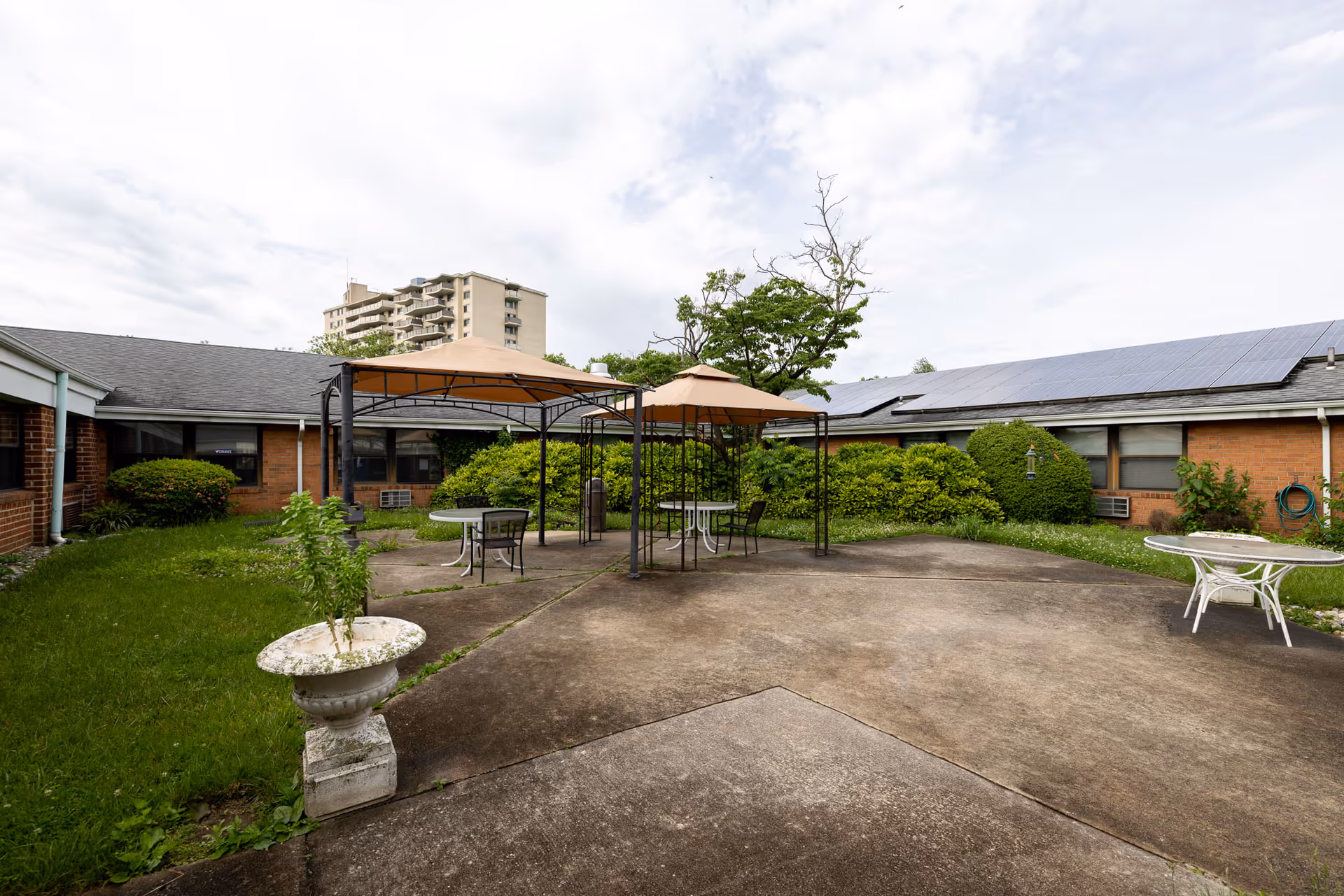 Outdoor courtyard area with concrete ground, two metal-framed gazebos with beige canopies, round tables and chairs. Surrounding the courtyard are low brick buildings with windows, bushes, and greenery. A tall apartment building is visible in the background under a partly cloudy sky.