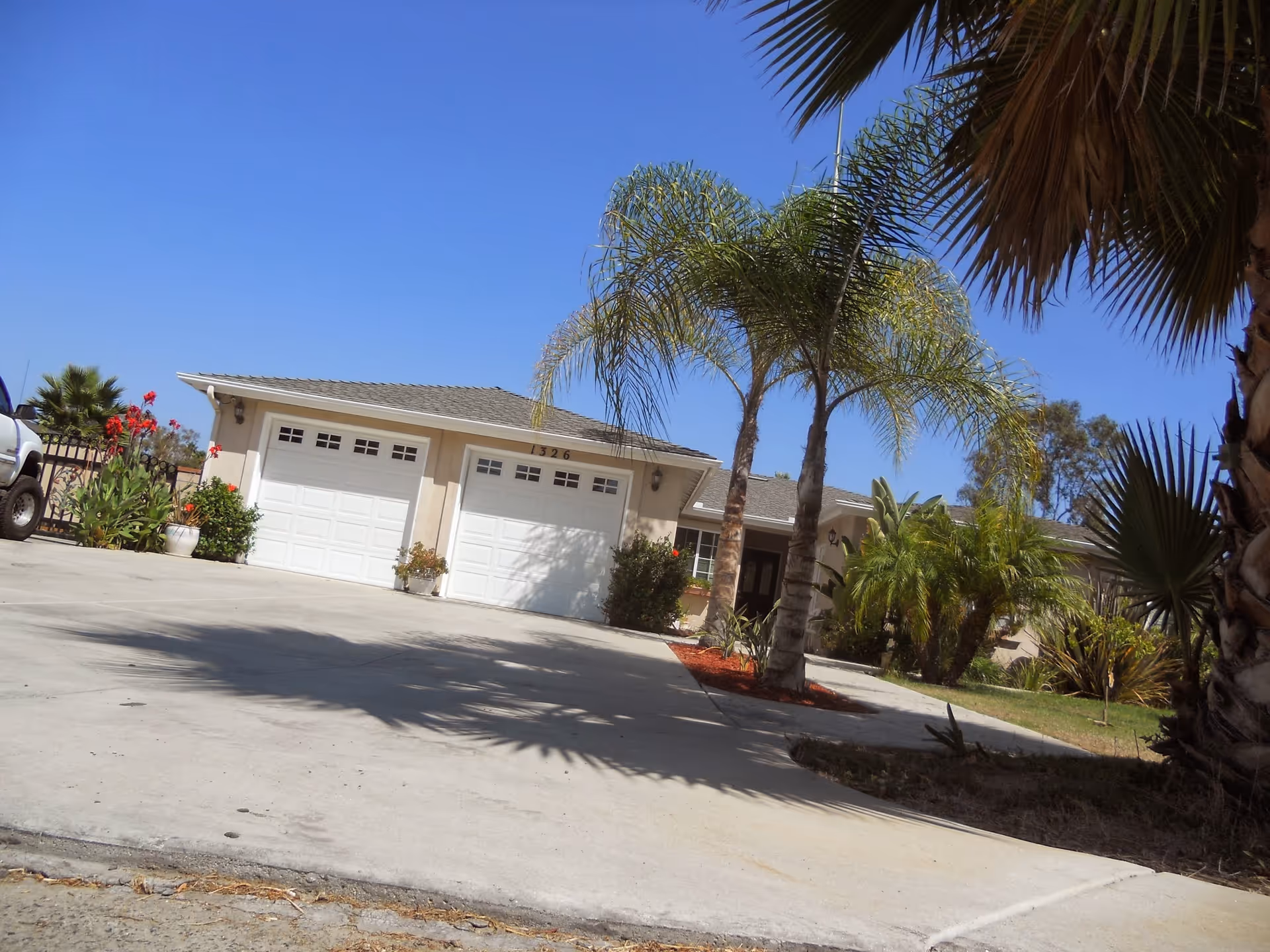 Exterior view of a single-story residential building with two white garage doors, surrounded by palm trees and other greenery under a clear blue sky.