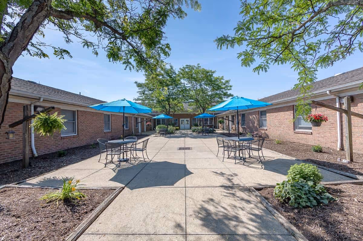 Sunny courtyard between single-story brick buildings with patio tables and bright blue umbrellas.