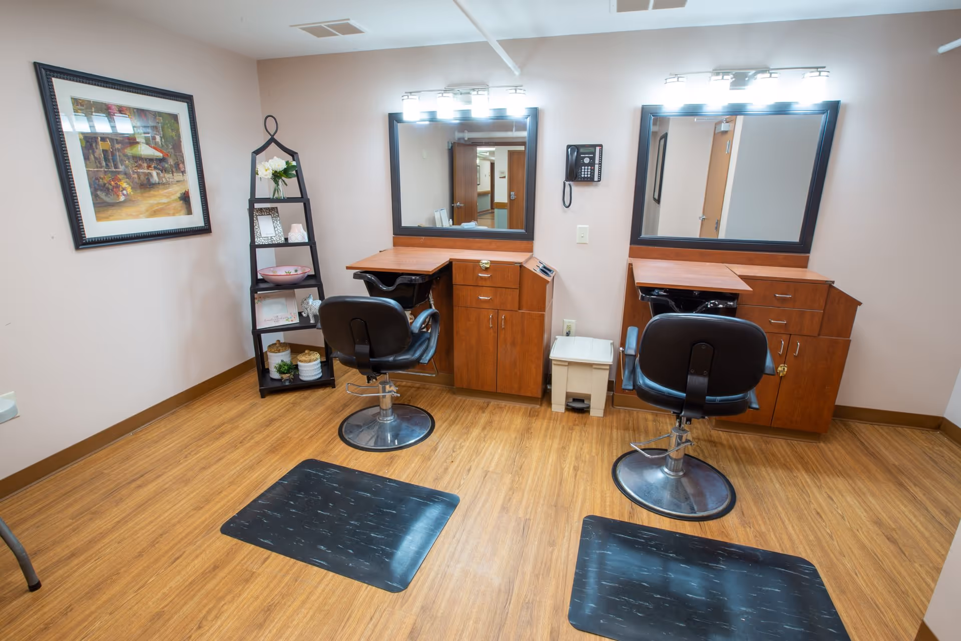 Small salon area with two styling stations, mirrors, black swivel chairs, shelving and wood flooring.