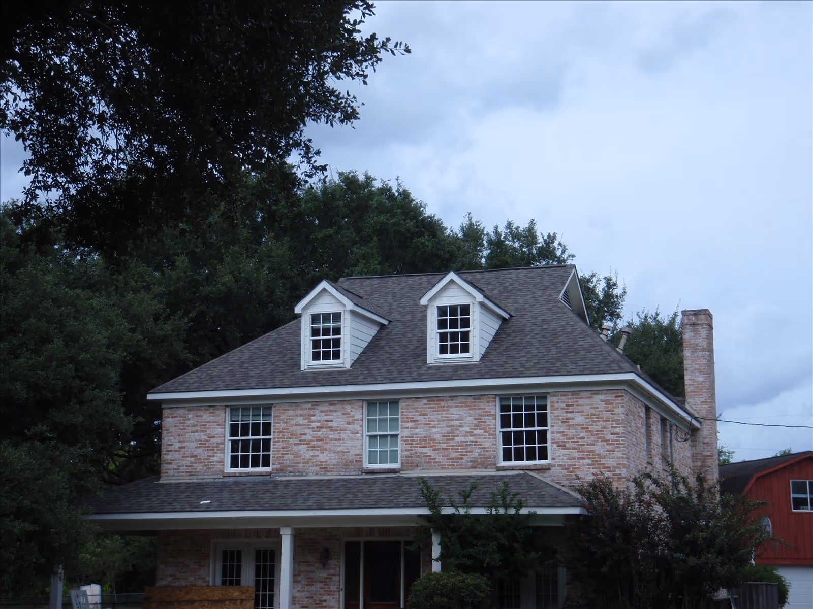 Front view of a two-story brick house with dormer windows, a covered porch, and surrounding trees under a cloudy sky.