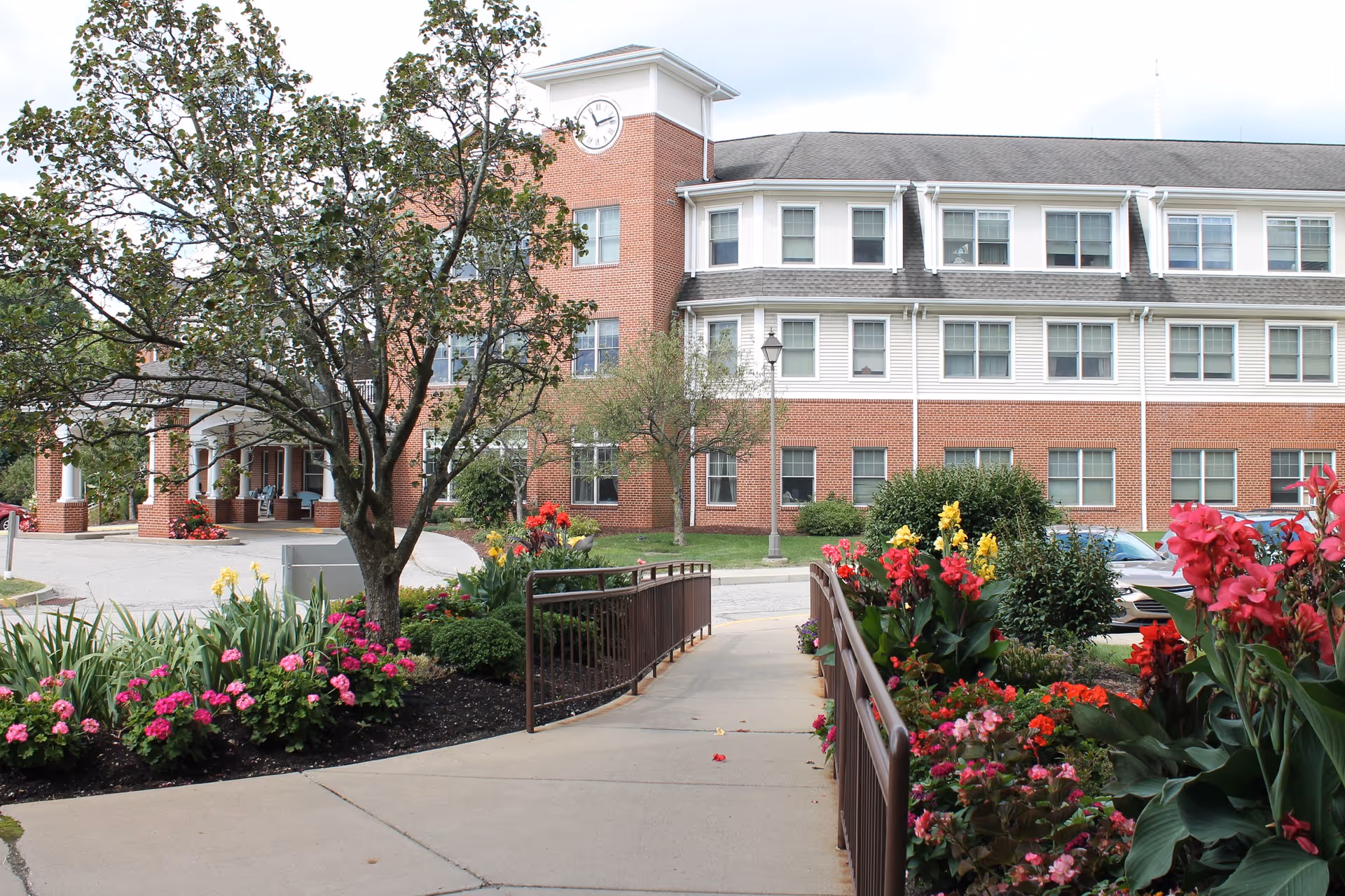 Front entrance and facade of a brick senior living building with a landscaped walkway and colorful flower beds.
