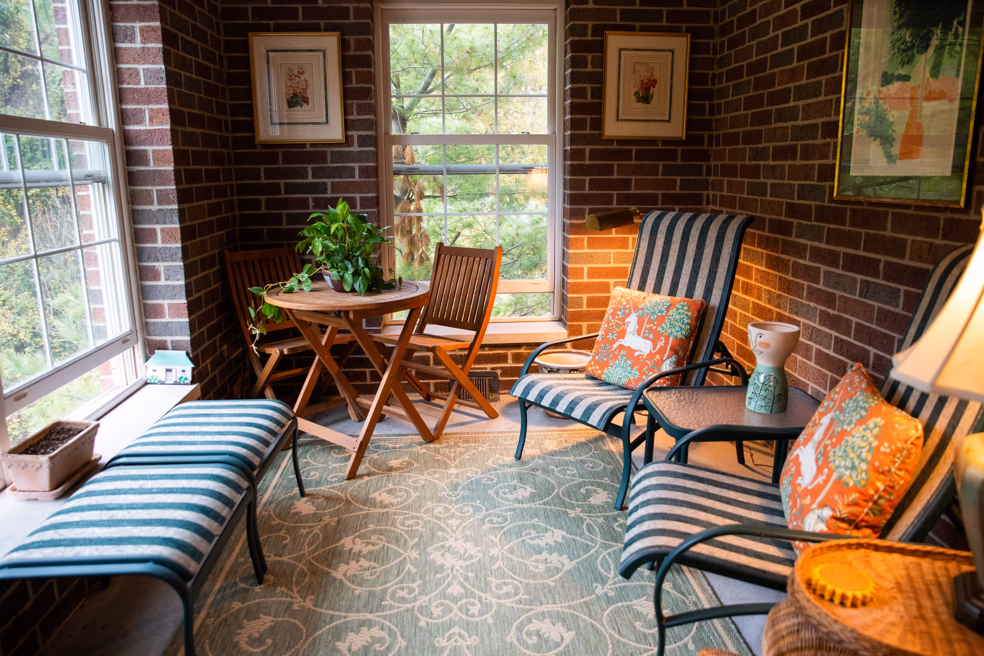 A cozy sunroom with brick walls and large windows showing greenery outside. The room features two striped cushioned chairs with orange decorative pillows, a small glass-top side table with a ceramic vase, a wooden round table with two wooden chairs, and a striped cushioned bench. There are framed floral artworks on the walls and a patterned area rug on the floor.