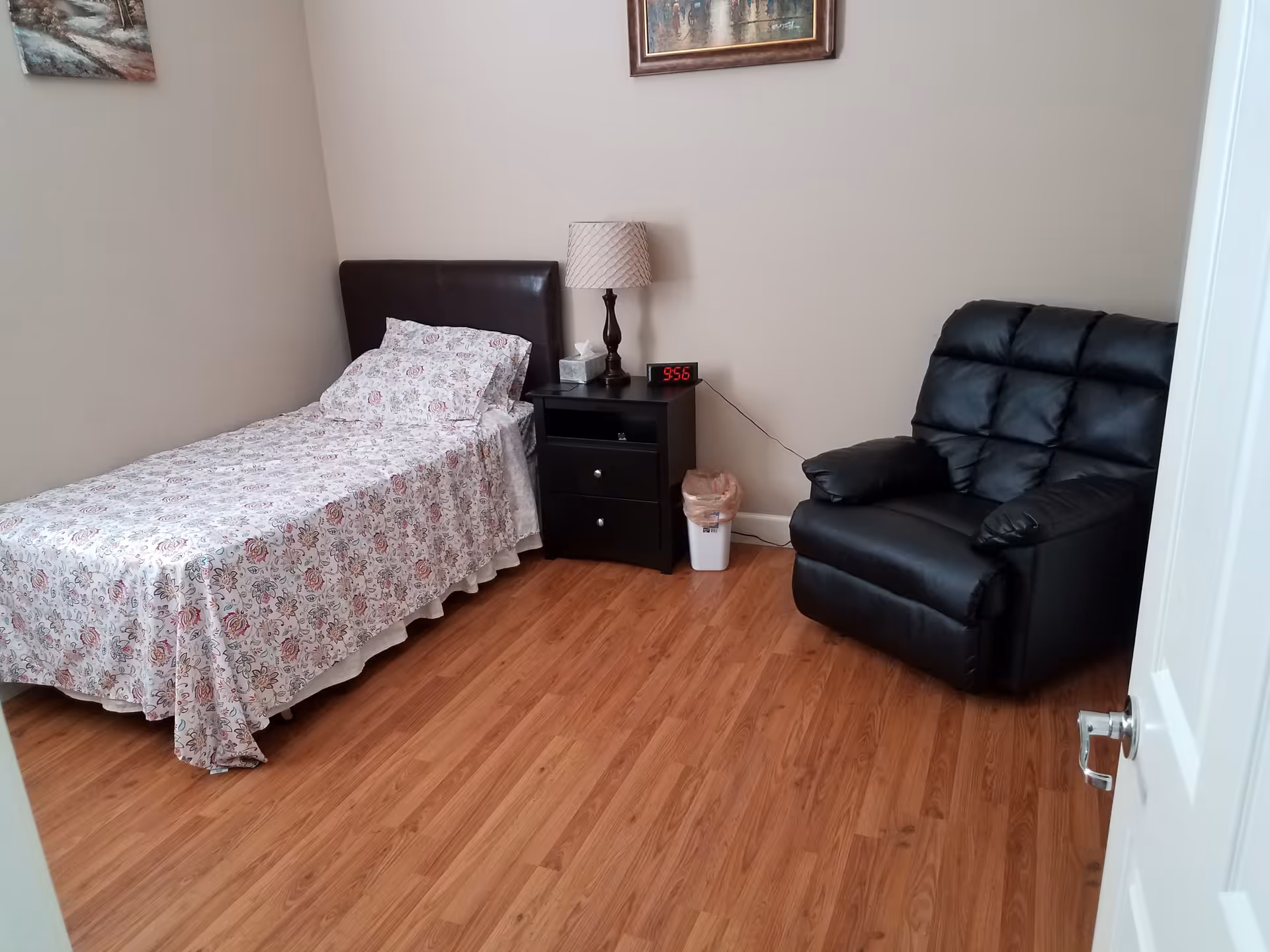 A small bedroom with a single bed covered in a floral bedspread, a black nightstand with a lamp, digital clock, and tissue box on it, a small trash can beside the nightstand, and a black recliner chair. The floor is wooden, and there are two framed pictures hanging on the beige walls.