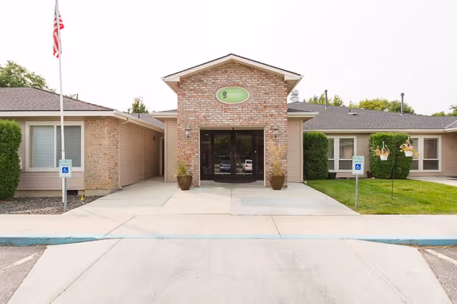 Front exterior view of Ashley Manor Senior Living building with a brick entrance, glass double doors, two potted plants on either side of the entrance, an American flag on the left, and handicap parking signs on both sides of the walkway.