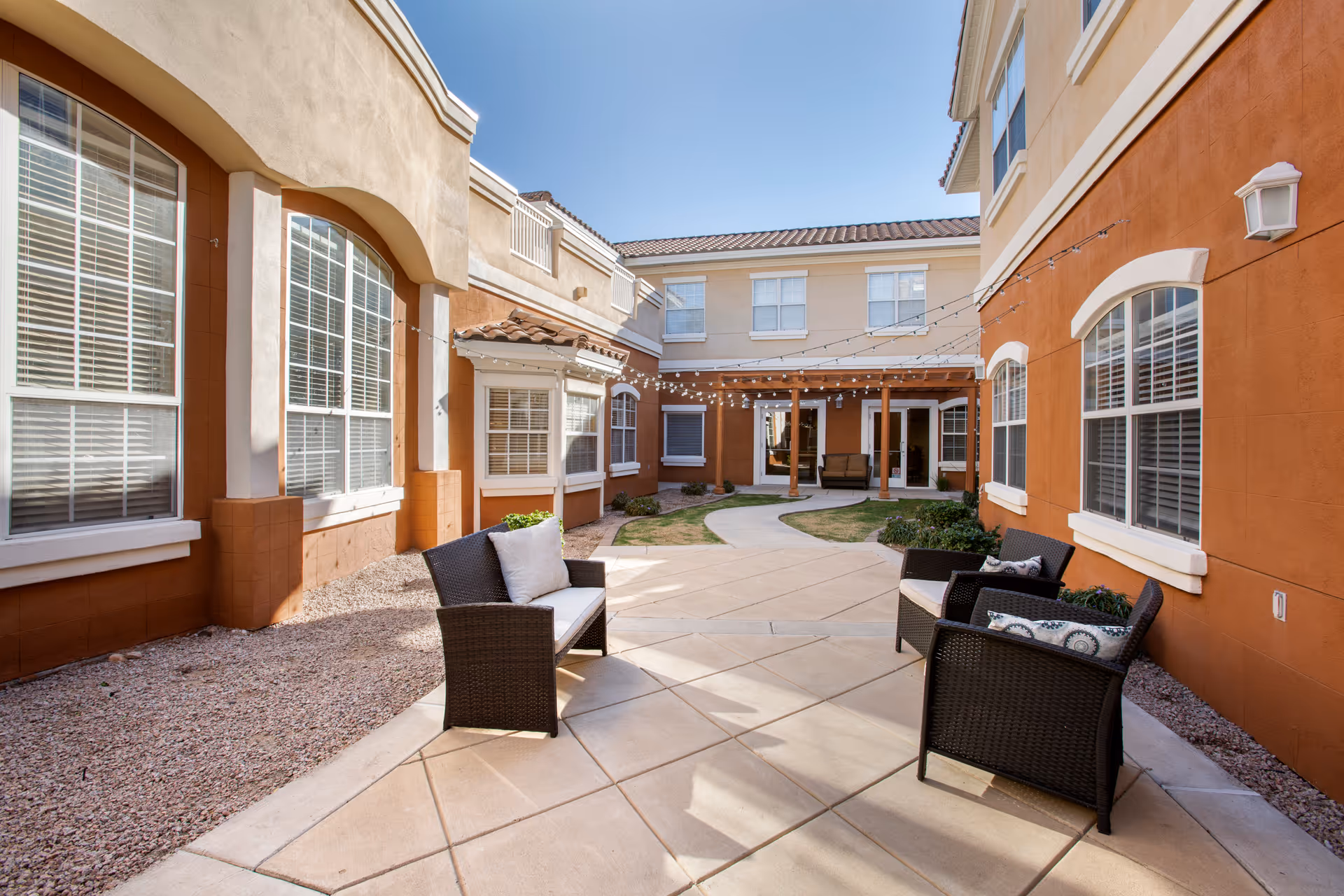 Enclosed sunny courtyard with wicker patio seating, paved walkways, landscaping, and string lights between two-story buildings.