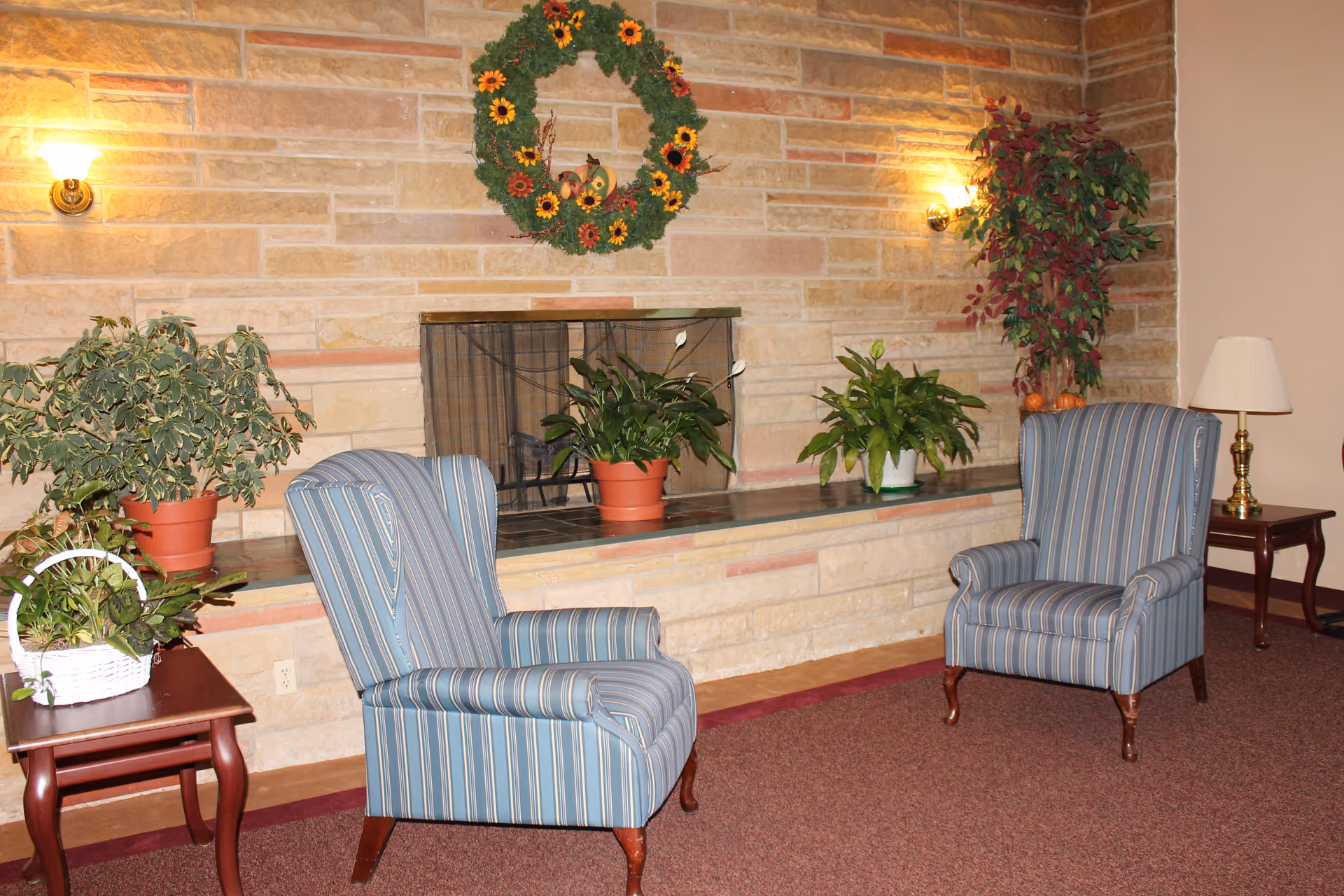 A cozy sitting area with two blue striped armchairs facing each other in front of a stone fireplace. The fireplace mantel is decorated with potted green plants and a wreath with sunflowers hangs above it. There are two side tables, one with a white basket of plants and the other with a lamp and small decorative items. The walls are made of light-colored stone and the floor is carpeted in a reddish-brown color.