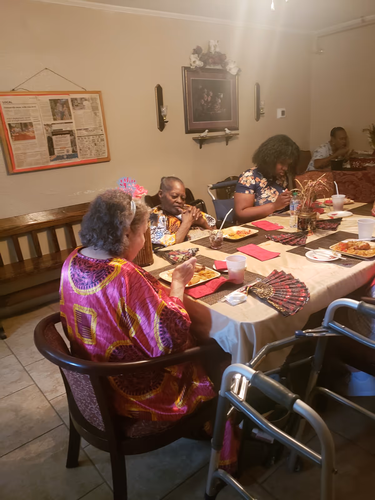 Several elderly individuals seated around a dining table in a room, eating a meal. The table is covered with a beige tablecloth and decorated with red napkins and a centerpiece. A walker is visible near the table, and the walls have framed pictures and a bulletin board.