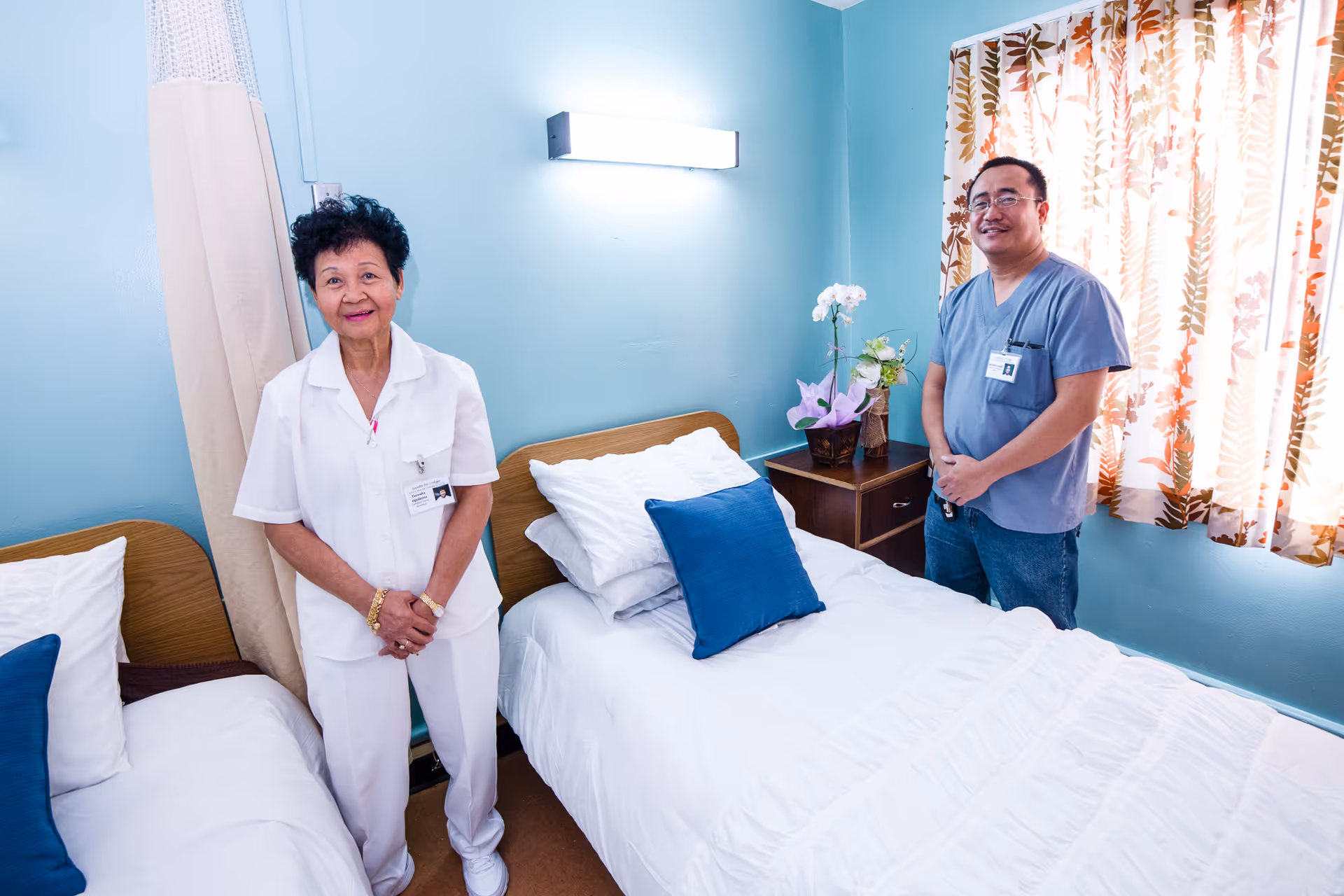 Two healthcare workers standing in a small bedroom with two single beds. The room has light blue walls, a window with floral curtains, and a nightstand with a flower arrangement between the beds. Both individuals are wearing name badges and smiling at the camera.