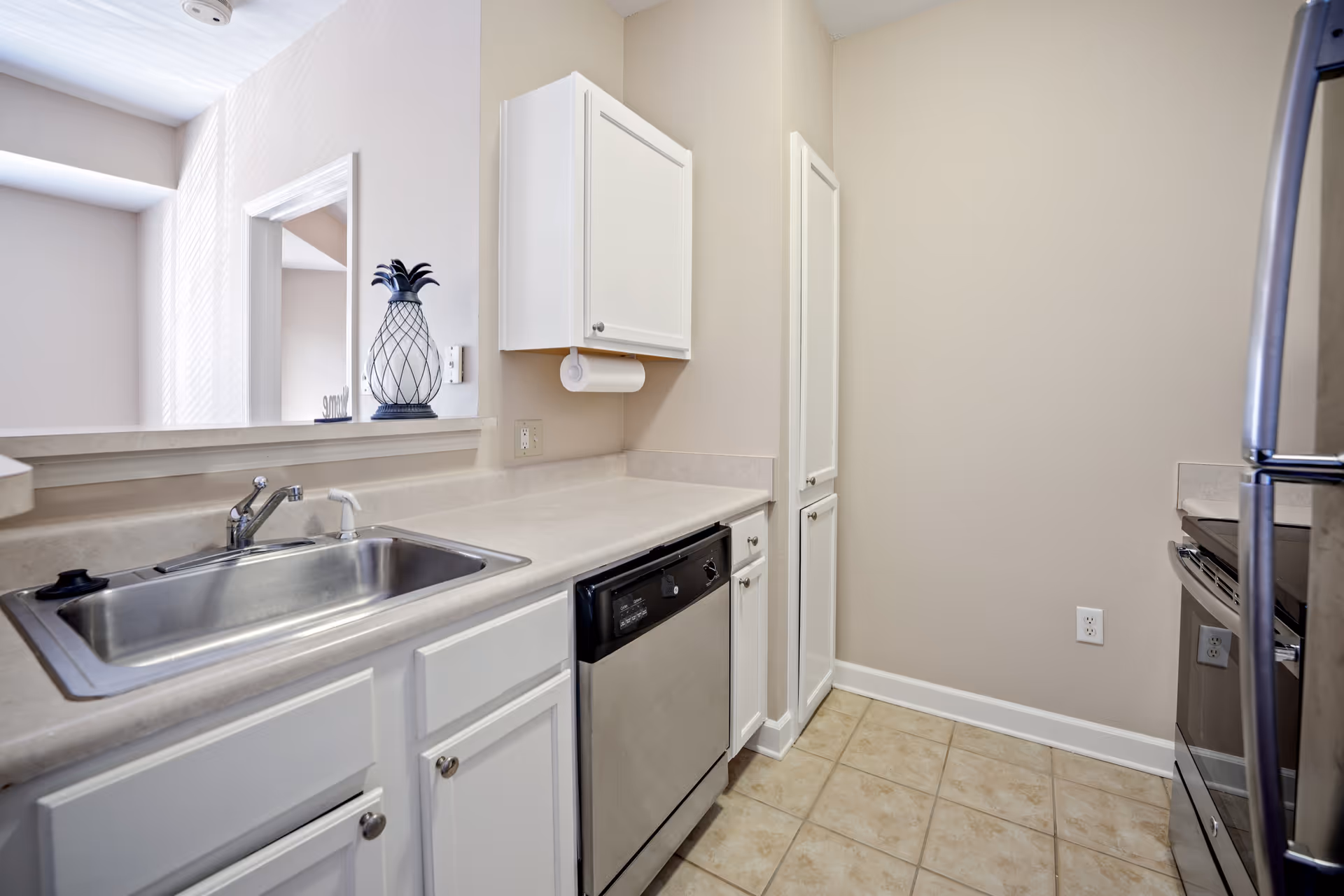 Small galley kitchen with a stainless steel sink and dishwasher, white cabinets, tile floor and a pass-through window.