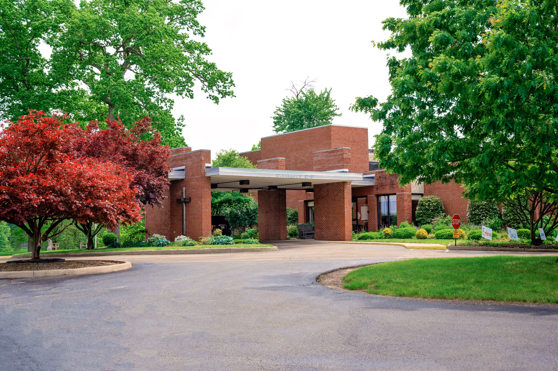 Exterior view of a brick building with a covered entrance driveway surrounded by green trees and landscaping, including a red-leaved tree on the left and a green-leaved tree on the right.