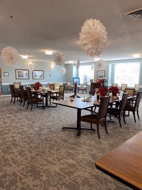 A spacious dining room with multiple tables and chairs arranged neatly. The tables are decorated with red poinsettia plants and place settings. White paper snowflake decorations hang from the ceiling. The room has light blue walls, carpeted floors with a patterned design, and several framed pictures on the walls. Large windows allow natural light to brighten the space.