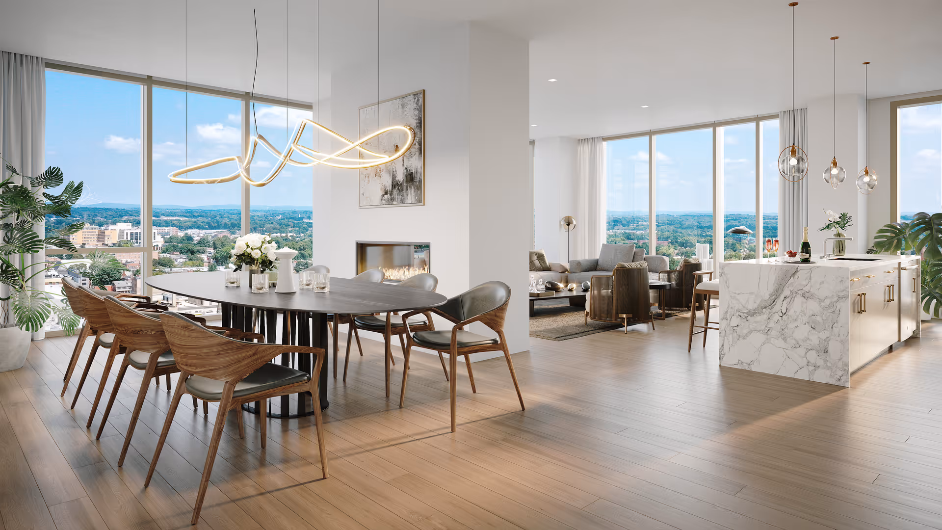 A modern open-concept living space with large floor-to-ceiling windows offering a panoramic view of a cityscape and distant hills. The space features a dining area with a dark oval table surrounded by wooden chairs with black cushions, a contemporary light fixture above the table, a fireplace built into a white wall, and a kitchen island with a white marble finish and pendant lights hanging above. The living area includes comfortable seating with sofas and armchairs, and the room is decorated with plants and minimalistic artwork.