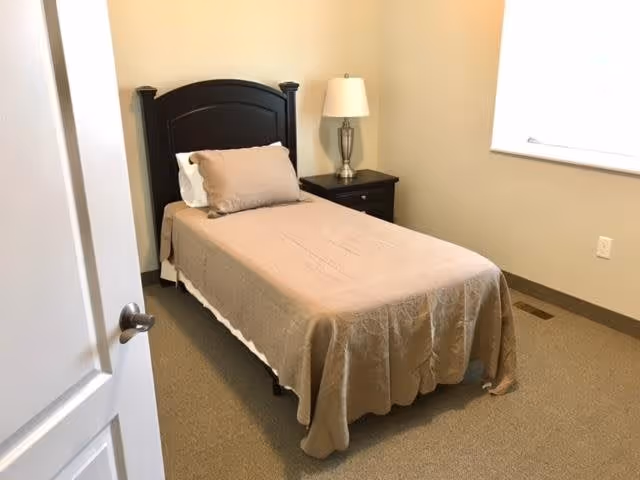 A small bedroom with a single bed covered in a beige bedspread and a matching pillow. Next to the bed is a dark wooden nightstand with a silver lamp on it. The room has beige walls, a window with closed blinds, and a beige carpeted floor. The door to the room is partially open.