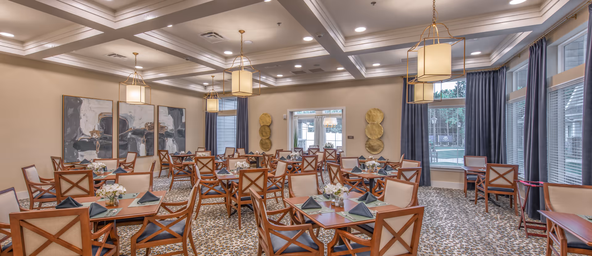 Bright, elegantly furnished dining room with multiple set tables and wooden chairs beneath decorative pendant lights and large windows.