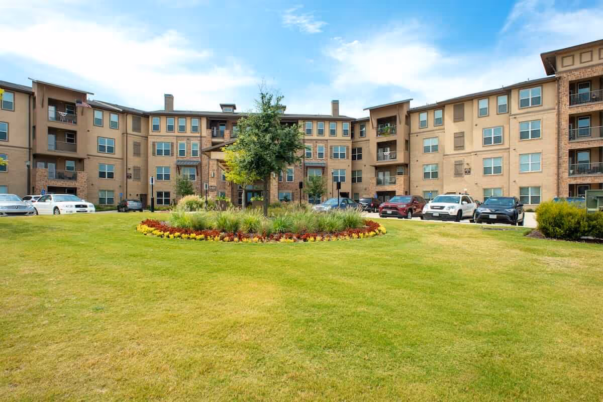 Exterior view of a multi-story senior living facility building with a well-maintained lawn and a circular flower bed with a tree in the center. Several parked cars are visible along the driveway in front of the building under a partly cloudy sky.
