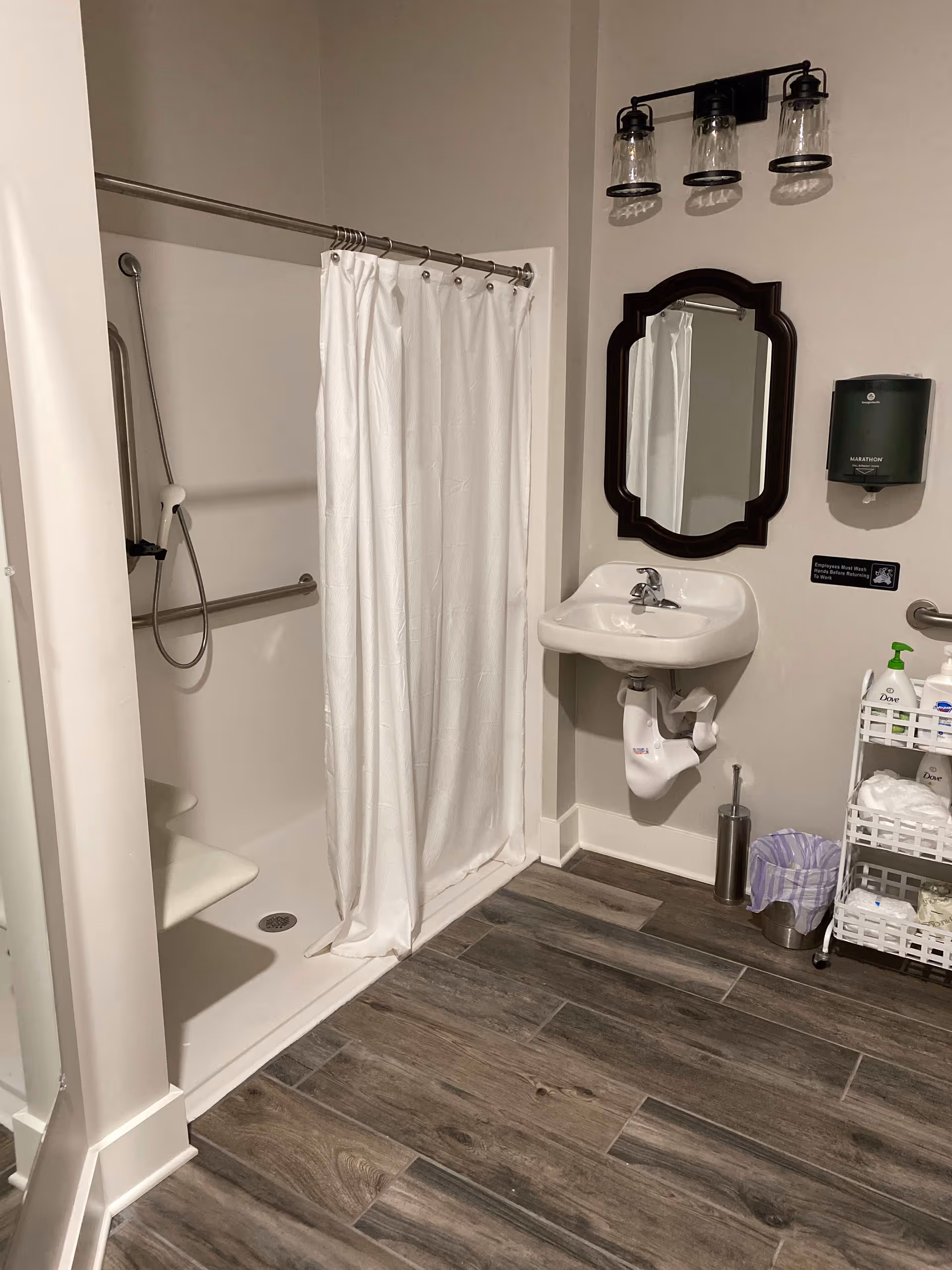 A clean and accessible bathroom featuring a walk-in shower with a white curtain, a wall-mounted sink with a mirror above it, a soap dispenser, a small trash can, and a white storage cart holding toiletries and towels. The floor has wood-like tiles.