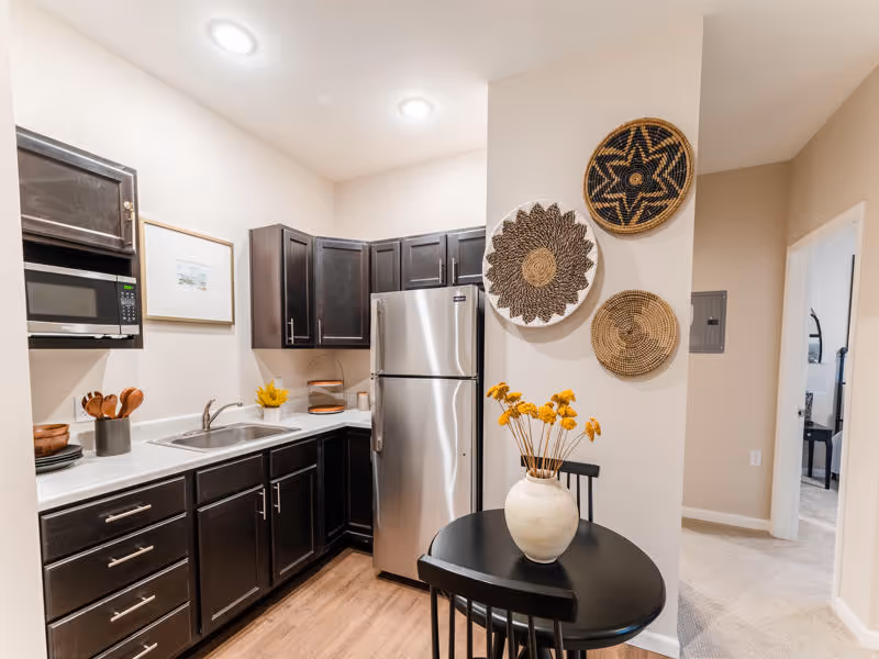 Small kitchenette with dark cabinets, stainless refrigerator, sink, and a round table with a vase of yellow flowers and woven wall baskets.