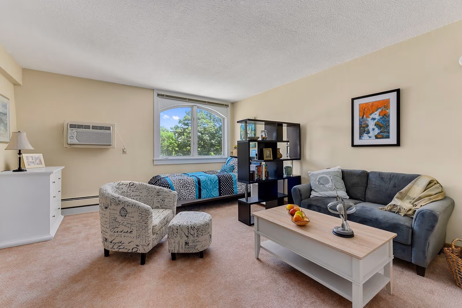 A cozy living space with a single bed near a window showing green trees outside. The room features a patterned armchair with matching ottoman, a gray sofa with a pillow and throw blanket, a white coffee table with decorative items and a bowl of fruit, a black open shelving unit with books and decor, a white dresser with a lamp and framed photos, and a wall-mounted air conditioner.
