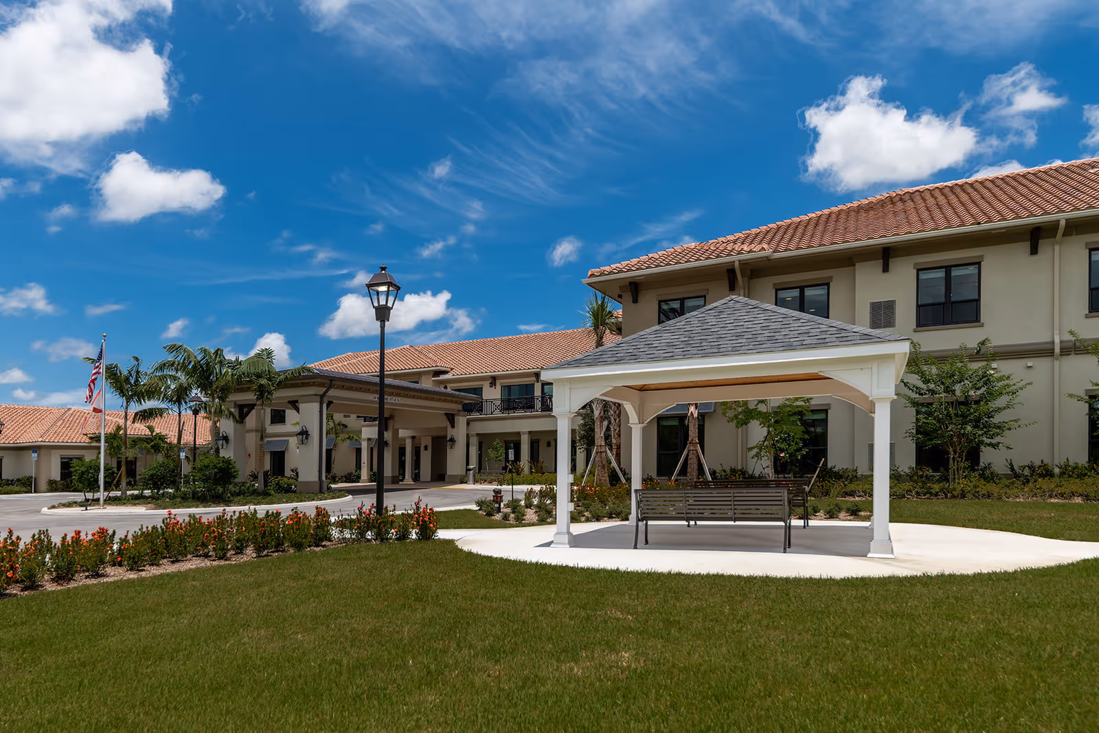 Front exterior of a senior living building with a white gazebo and bench, landscaped lawn, and tiled-roof entrance under a blue sky.