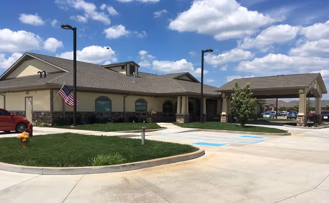 Exterior view of a single-story building with beige walls and stone accents, featuring a covered entrance and an American flag on a pole near the sidewalk. The building is surrounded by a paved driveway, green grass, and a few small trees under a blue sky with scattered clouds.