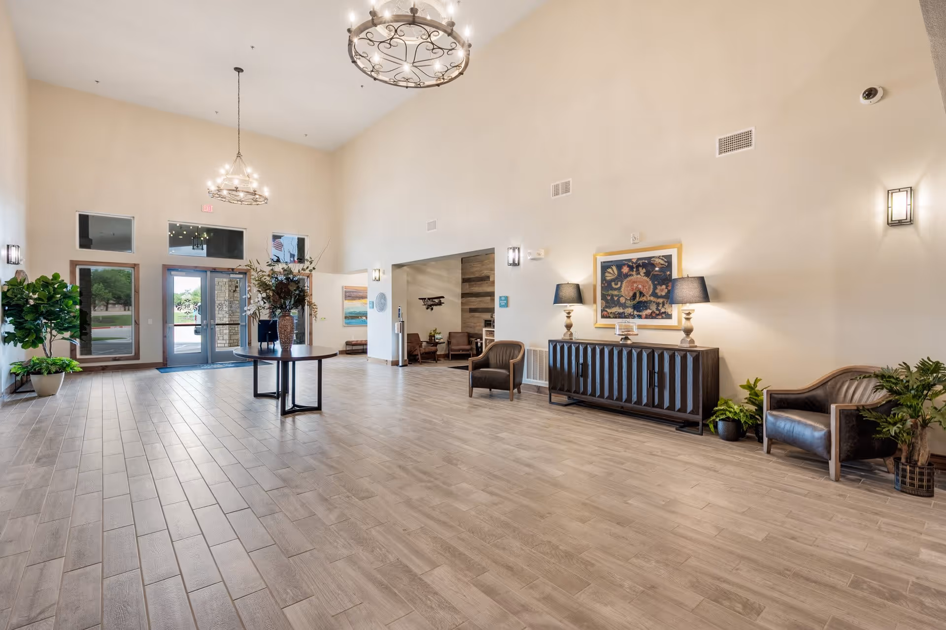 Spacious and well-lit senior living facility lobby with high ceilings, two large chandeliers, a round table with a floral arrangement in the center, comfortable chairs, a wooden sideboard with lamps, framed artwork on the walls, and several potted plants. The entrance door and windows allow natural light to enter the room.