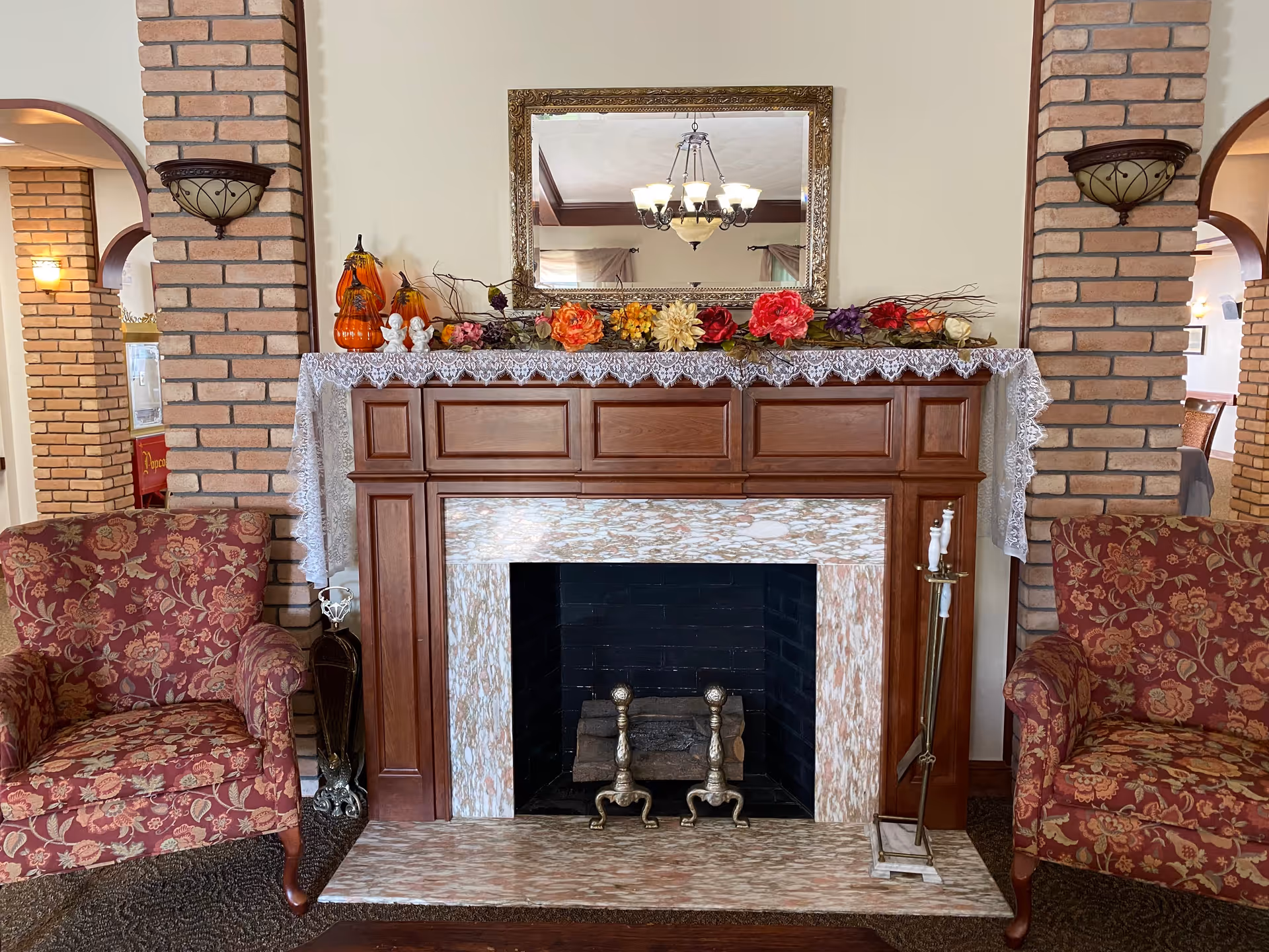 A cozy living room area featuring a decorative fireplace with a wooden mantel adorned with lace cloth, artificial flowers, and small pumpkin decorations. Two floral upholstered armchairs flank the fireplace, and a large ornate mirror hangs above it. The room has brick columns and warm lighting fixtures.