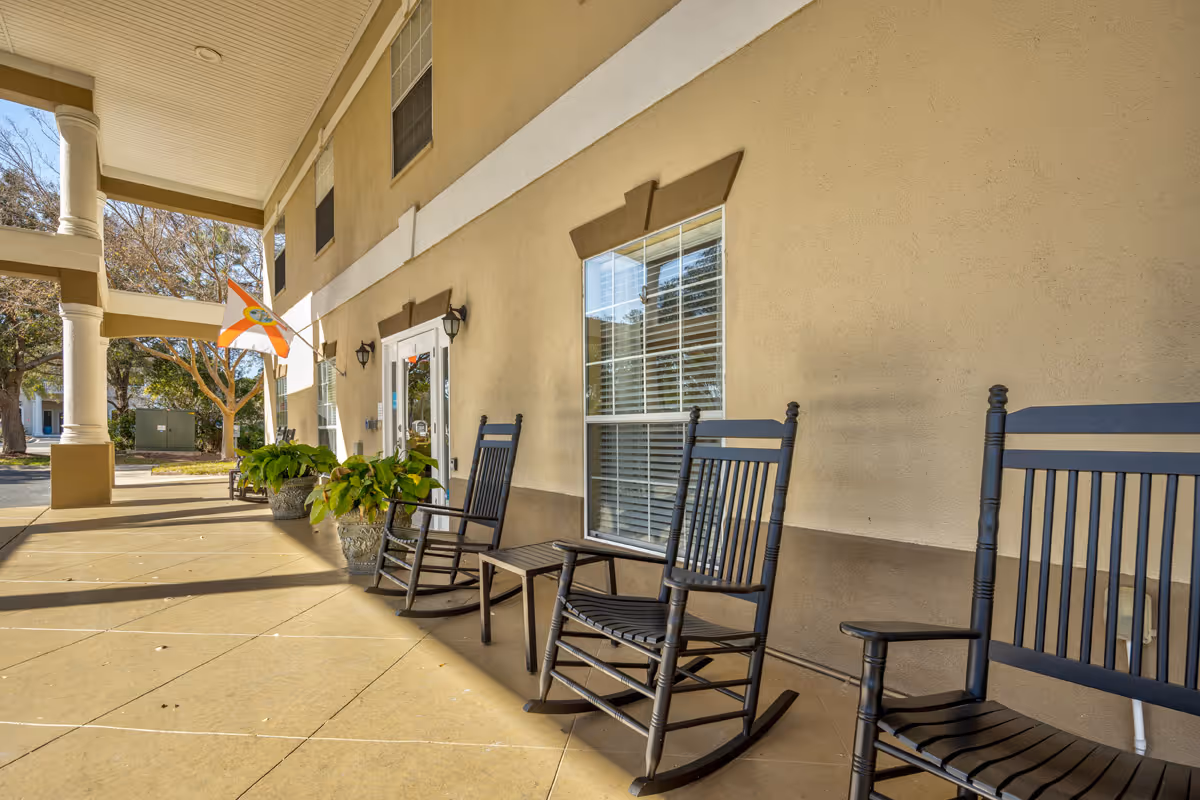 Covered outdoor patio area with several black rocking chairs and potted plants along the wall of a beige building. A flag is mounted near the entrance door, and trees are visible in the background.