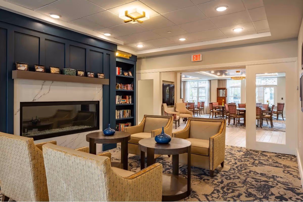 A cozy sitting area with beige armchairs and round wooden tables, each table decorated with a blue vase. The room features a modern fireplace set in a dark blue paneled wall with a marble surround, and a built-in bookshelf filled with books and framed photos. Beyond the sitting area, there is a dining room with multiple tables and chairs, large windows letting in natural light, and an exit sign above the doorway.