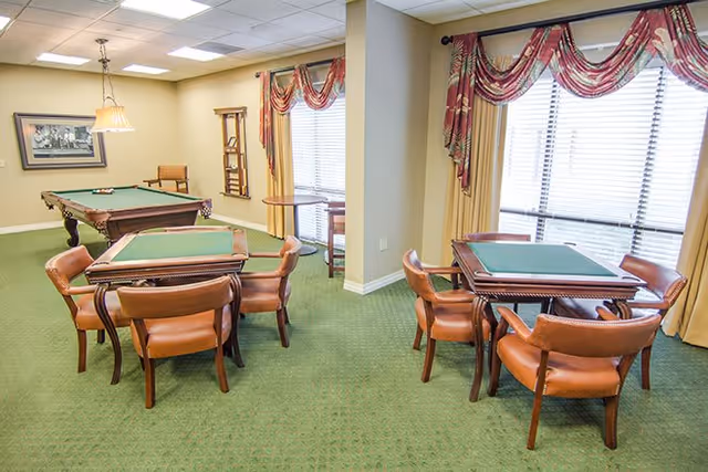 A well-lit recreational room with green carpet featuring two card tables with green felt tops surrounded by brown leather chairs. In the background, there is a pool table with a green felt surface. The room has large windows with red and gold drapes, beige walls, and a framed picture on the wall.