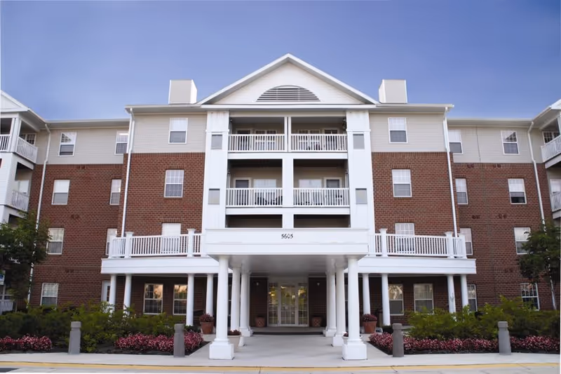 Front exterior view of Marwood Senior Apartments, a multi-story building with a combination of red brick and light-colored siding. The entrance features white columns supporting a covered porch area, balconies with white railings, and well-maintained landscaping with bushes and flowers along the walkway.