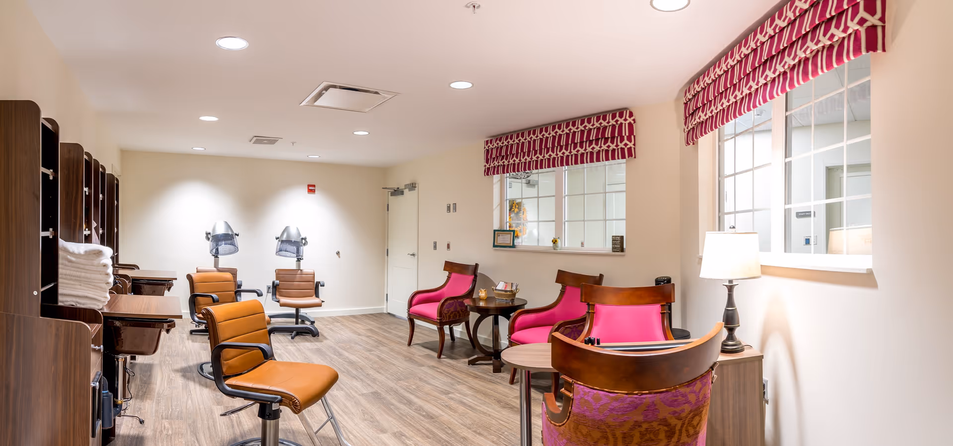 Interior view of a salon area in a senior living facility with brown salon chairs and hair drying stations on the left, and a seating area with pink cushioned chairs and small tables on the right. The room has wood flooring, white walls, and windows with red and white patterned valances.