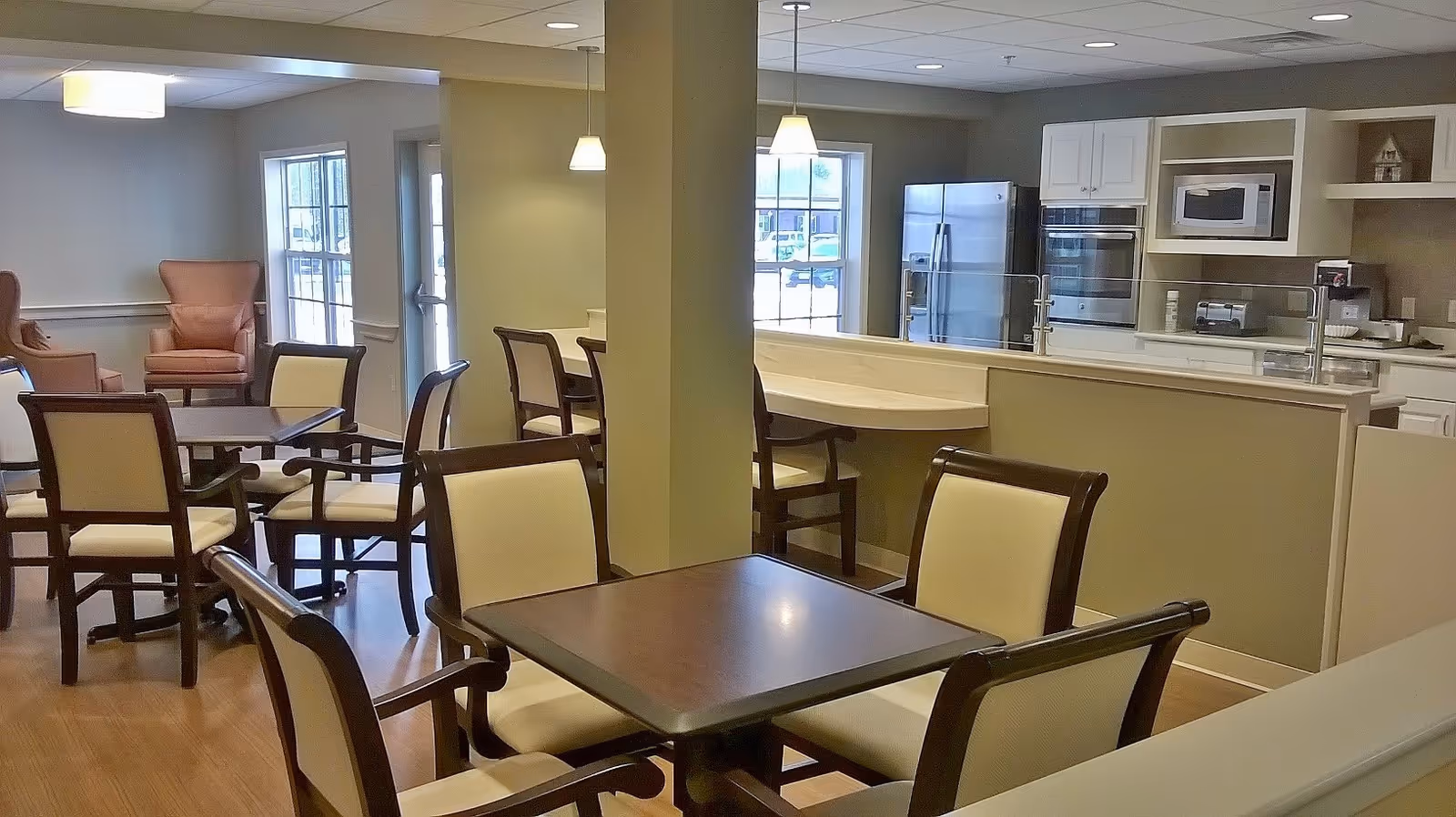 Interior view of a senior living facility dining area with several tables and chairs arranged neatly. The room has large windows letting in natural light, and in the background, there is a kitchen area with white cabinets, a refrigerator, microwave, and oven. The flooring is wooden, and the walls are painted in neutral tones.