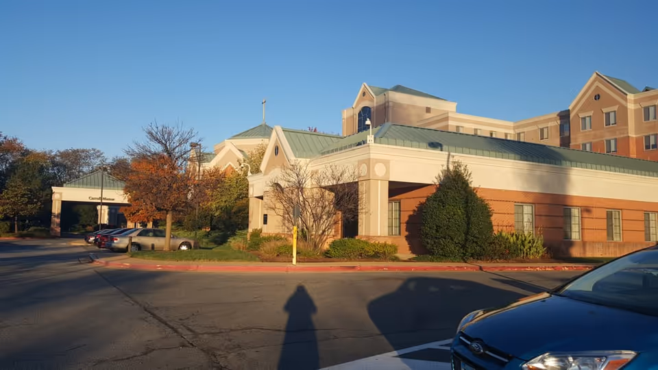 Exterior view of Carroll Manor Nursing & Rehab facility showing a multi-story building with a green roof, brick and beige walls, surrounded by trees and parked cars under a clear blue sky.