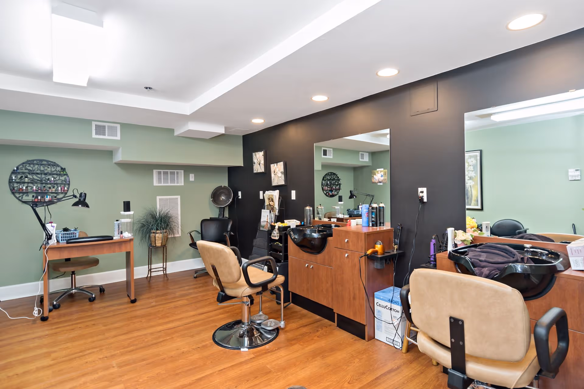 Salon-style interior with styling chairs, shampoo sinks, mirrors, and a manicure desk on wood flooring.