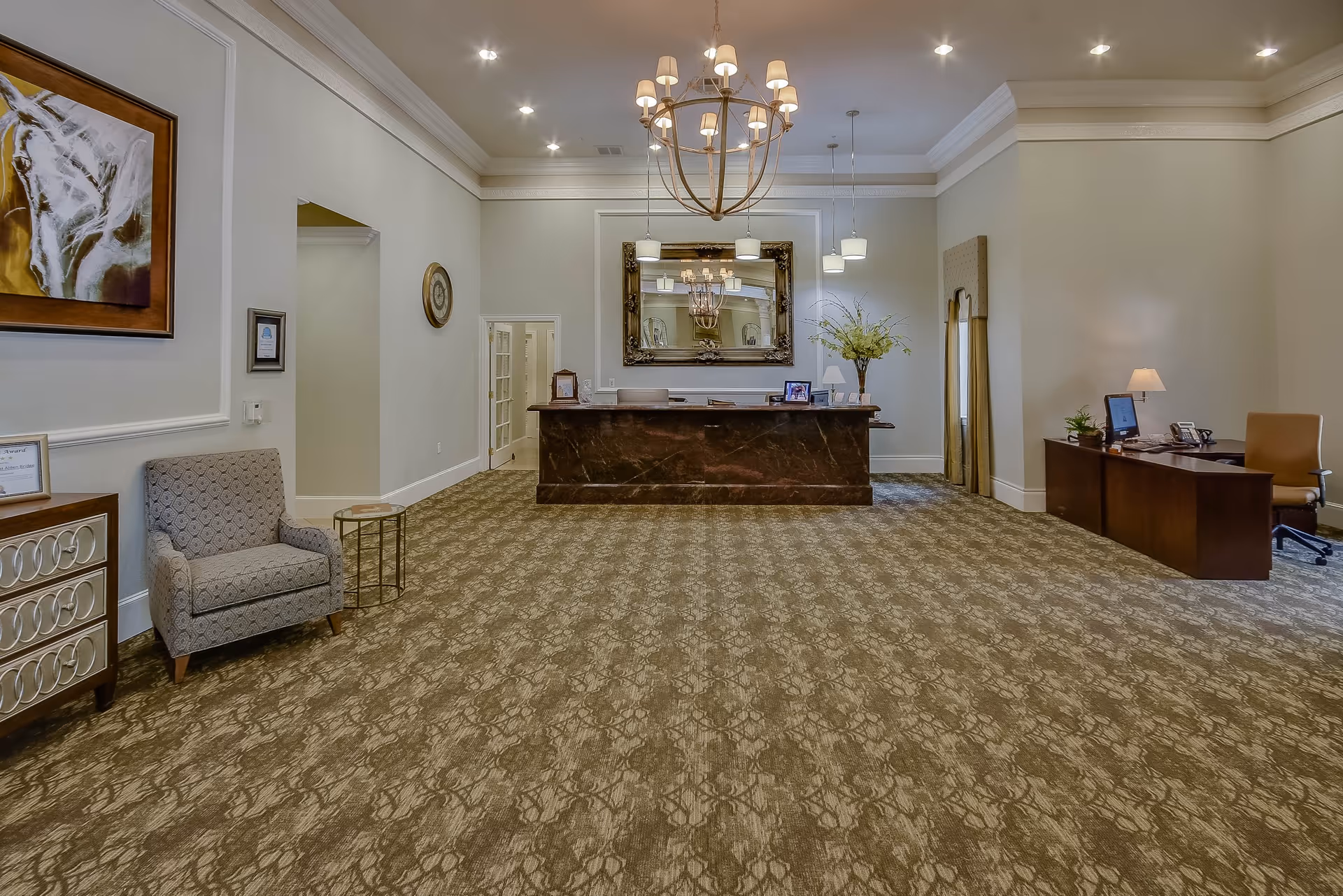 A spacious reception area with a patterned carpet, a large chandelier hanging from the ceiling, and a dark marble front desk with a large ornate mirror behind it. To the left, there is a gray upholstered armchair next to a small round side table and a decorative cabinet. On the right side, there is a wooden desk with a computer, lamp, and office chair. The walls are light-colored with crown molding and some framed artwork and decorations.